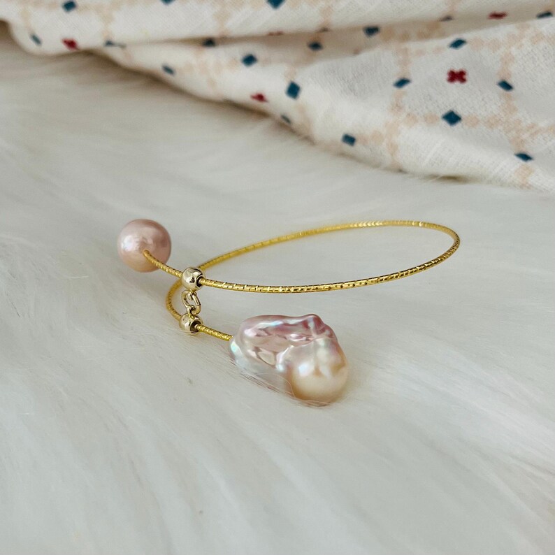 Peut inclure: Un bracelet jonc en or avec deux perles baroques roses. Le bracelet est fait de fil d'or fin et textur&eacute;.