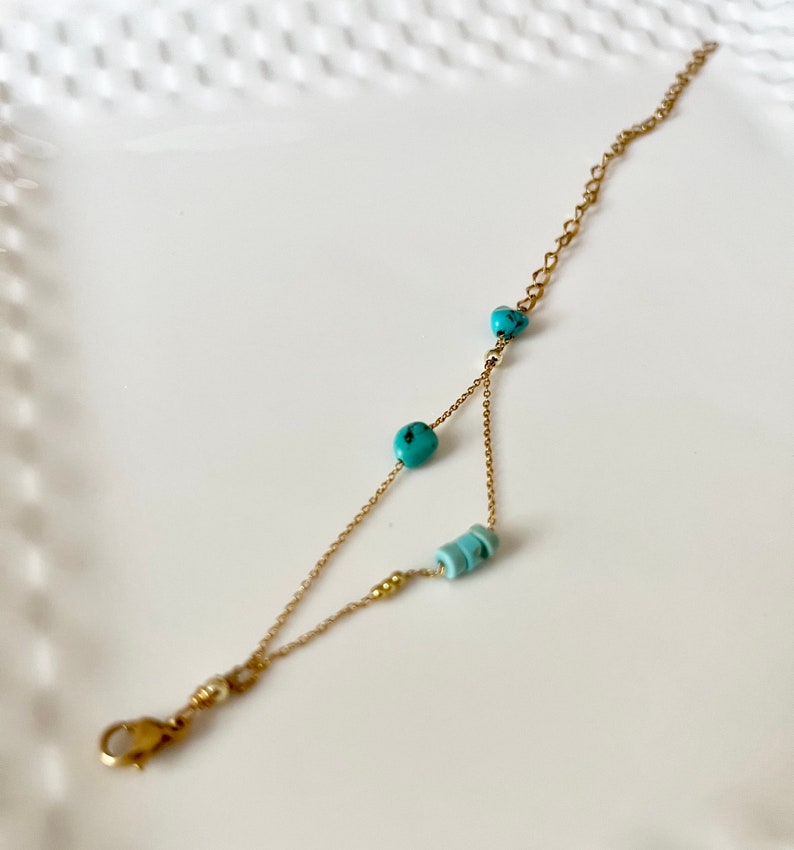 May include: A delicate gold chain bracelet with turquoise and blue beads. The bracelet has a lobster clasp closure.