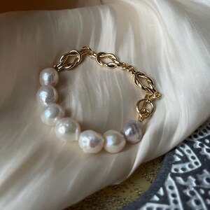 Puede incluir: Un brazalete de cadena dorada con un dise&ntilde;o de nudo y una hebra de perlas blancas.