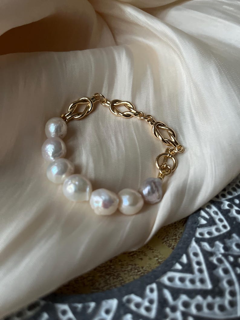 Puede incluir: Un brazalete de cadena dorada con un dise&ntilde;o de nudo y una hebra de perlas blancas.
