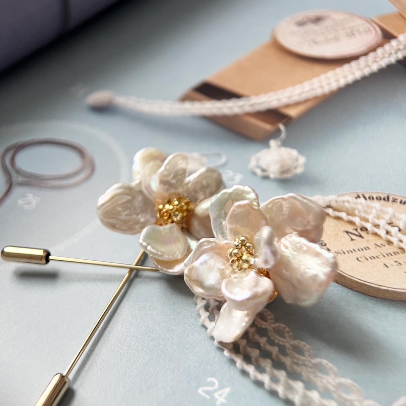 Puede incluir: Un par de broches de flores de perlas blancas con detalles dorados. Las broches est&aacute;n sujetas a un pasador de seguridad dorado.