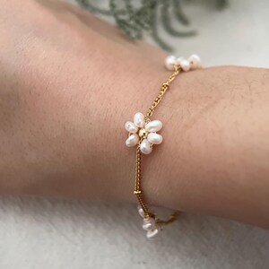 May include: A delicate gold chain bracelet with small white pearl flowers. The bracelet is on a person's wrist.