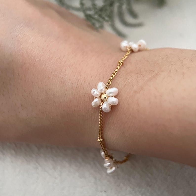 May include: A delicate gold chain bracelet with small white pearl flowers. The bracelet is on a person's wrist.