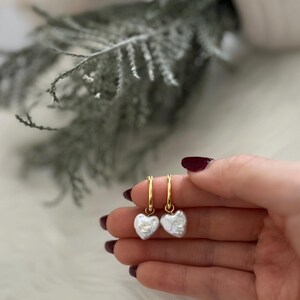 Op de afbeelding: Een paar gouden creolen met een witte parelhart-hanger aan elke oorring.