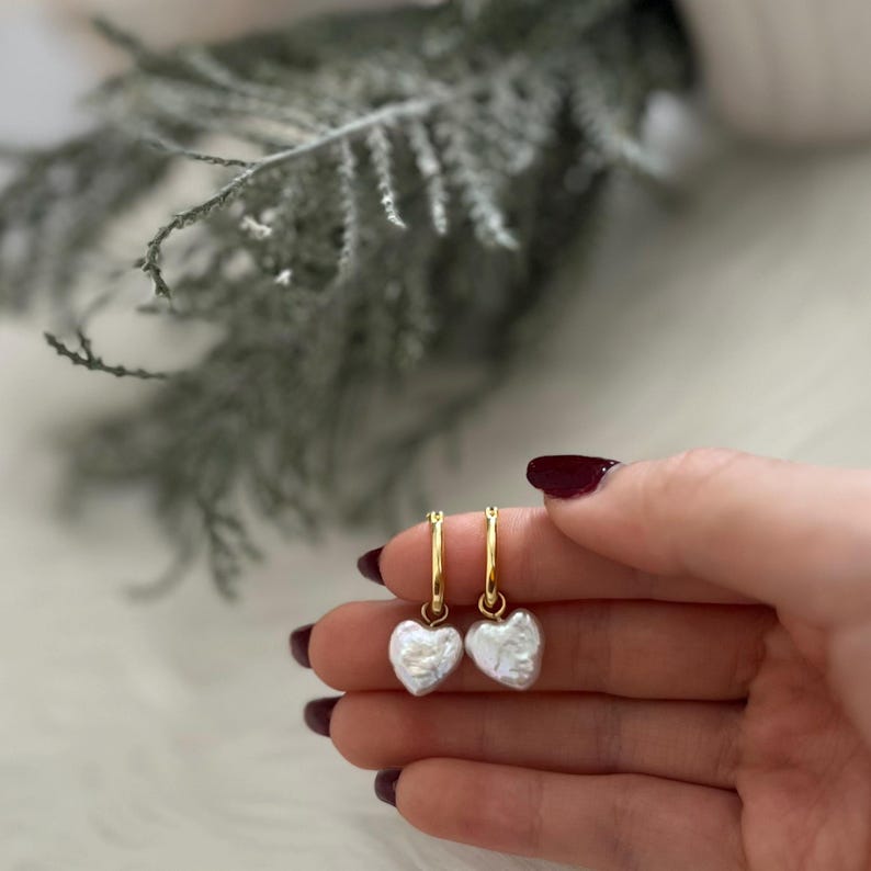 Op de afbeelding: Een paar gouden creolen met een witte parelhart-hanger aan elke oorring.