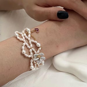 May include: A multi-strand pearl bracelet with a gold chain and a small red heart charm. The bracelet features a flower design with white petals and gold accents. The pearls are white and vary in size.