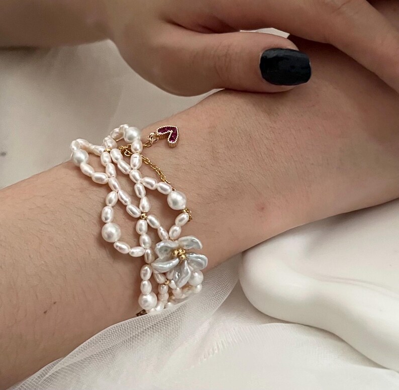 May include: A multi-strand pearl bracelet with a gold chain and a small red heart charm. The bracelet features a flower design with white petals and gold accents. The pearls are white and vary in size.
