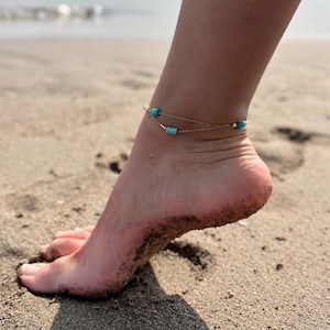 May include: A gold anklet with turquoise beads on a woman's ankle. The anklet is on a sandy beach.