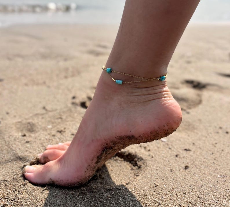 May include: A gold anklet with turquoise beads on a woman's ankle. The anklet is on a sandy beach.