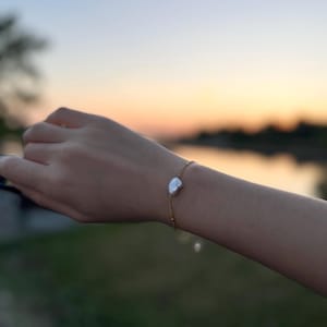 Puede incluir: Una delicada pulsera dorada con una sola perla blanca de forma irregular. La pulsera se muestra en una muñeca sobre un fondo borroso de una puesta de sol sobre el agua. La cadena de oro es fina y elegante.