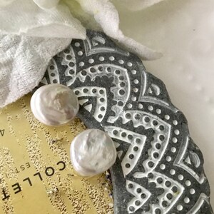 May include: Two white pearl earrings on a gray and white patterned surface. The earrings are round and have a smooth, glossy finish.