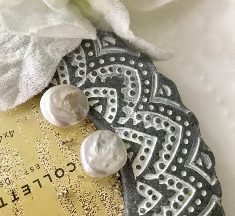 May include: Two white pearl earrings on a gray and white patterned surface. The earrings are round and have a smooth, glossy finish.