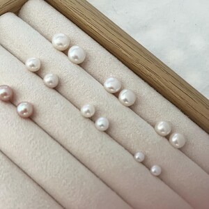 Peut inclure: Une collection de perles blanches et roses de diff&eacute;rentes tailles, dispos&eacute;es dans un pr&eacute;sentoir &agrave; bijoux beige clair avec un cadre en bois. Les perles sont rondes et ont un &eacute;clat brillant, refl&eacute;tant la lumi&egrave;re. Le tissu doux du pr&eacute;sentoir offre un fond d&eacute;licat.