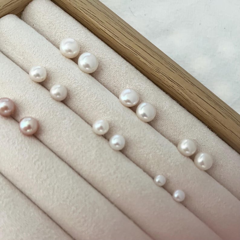Peut inclure: Une collection de perles blanches et roses de diff&eacute;rentes tailles, dispos&eacute;es dans un pr&eacute;sentoir &agrave; bijoux beige clair avec un cadre en bois. Les perles sont rondes et ont un &eacute;clat brillant, refl&eacute;tant la lumi&egrave;re. Le tissu doux du pr&eacute;sentoir offre un fond d&eacute;licat.
