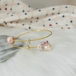 Peut inclure: Un bracelet jonc en or avec deux perles. Une perle est ronde et l'autre a une forme irr&eacute;guli&egrave;re. Le bracelet est sur une surface blanche et duveteuse.