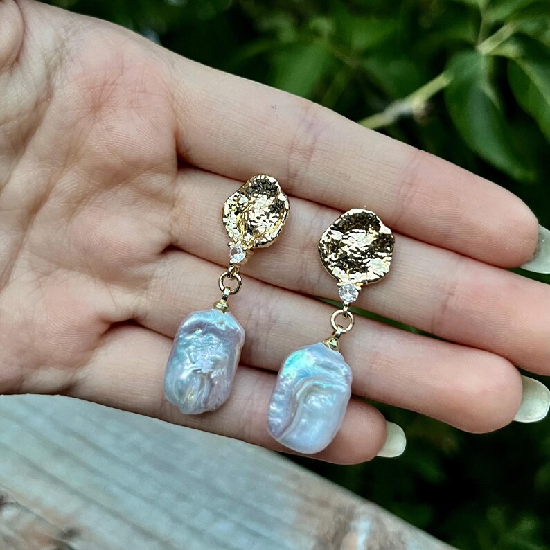 Puede incluir: Un par de pendientes de oro con un acabado texturizado y martillado. Cada pendiente presenta un peque&ntilde;o cristal transparente y una perla grande de forma irregular.