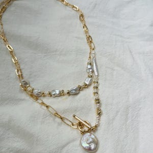 May include: A layered gold-tone necklace with freshwater pearls and a toggle clasp. The necklace has two strands, one with irregular pearls and the other with a delicate chain. The bottom strand has a large, iridescent pearl pendant.