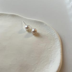 Peut inclure: Une paire de boucles d'oreilles &agrave; clous en perles, avec des tiges en argent, pos&eacute;es sur un plat en c&eacute;ramique blanc de forme organique. Les perles ont un &eacute;clat doux et iris&eacute;, refl&eacute;tant des nuances subtiles de rose et de cr&egrave;me. Bijou classique.