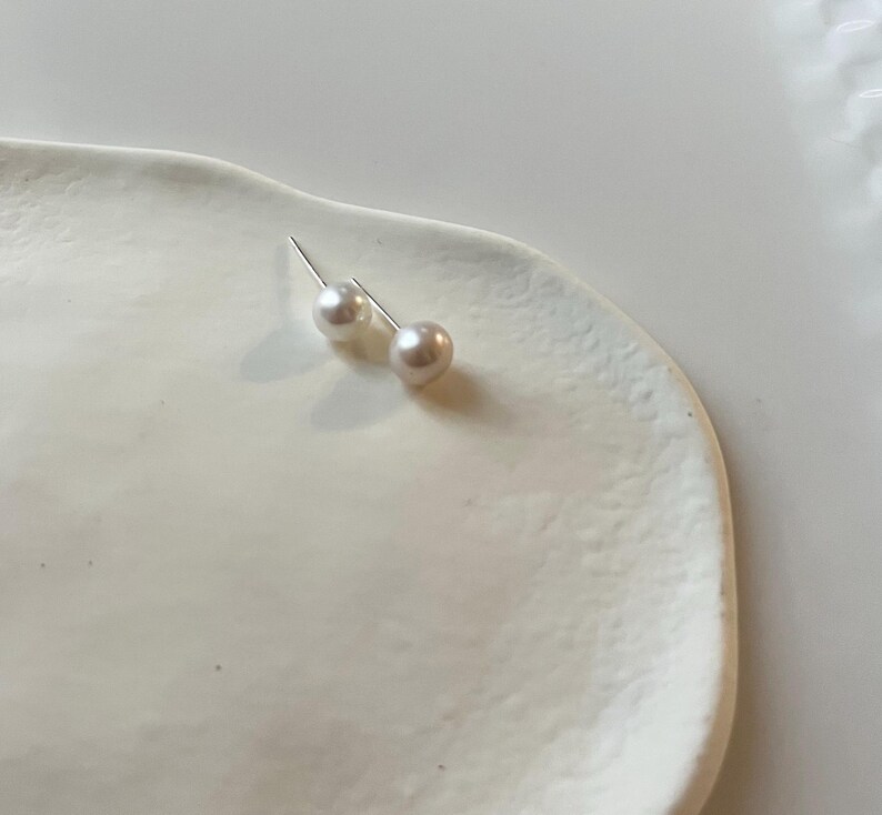 Peut inclure: Une paire de boucles d'oreilles &agrave; clous en perles, avec des tiges en argent, pos&eacute;es sur un plat en c&eacute;ramique blanc de forme organique. Les perles ont un &eacute;clat doux et iris&eacute;, refl&eacute;tant des nuances subtiles de rose et de cr&egrave;me. Bijou classique.