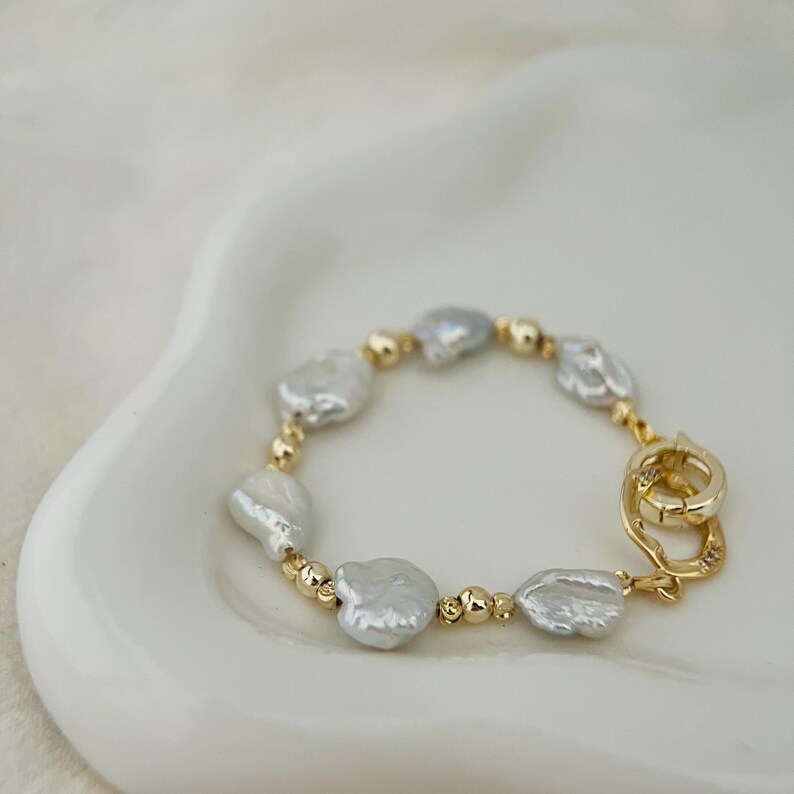 Peut inclure: Un bracelet dor&eacute; orn&eacute; de perles blanches de forme irr&eacute;guli&egrave;re et de petites perles dor&eacute;es. Le bracelet est dot&eacute; d'un fermoir dor&eacute;. Le bijou est pr&eacute;sent&eacute; sur une surface blanche et incurv&eacute;e.