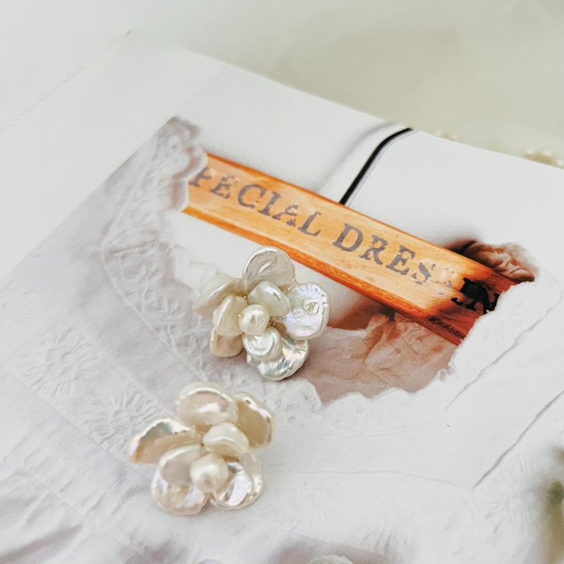 Op de afbeelding: Twee bloemvormige oorbellen met een parelcentrum en iriserende bloemblaadjes. De oorbellen liggen op een wit oppervlak met de tekst "SPECIAL DRESS" op een houten hanger. De oorbellen zijn waarschijnlijk voor een speciale gelegenheid.