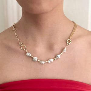 Peut inclure: Un collier dor&eacute; orn&eacute; de perles blanches irr&eacute;guli&egrave;res et de petites perles dor&eacute;es. Le collier a une cha&icirc;ne et une fermeture &agrave; fermoir d'un c&ocirc;t&eacute; et une cha&icirc;ne &agrave; plusieurs brins de l'autre. Le collier est port&eacute; sur un haut rouge.