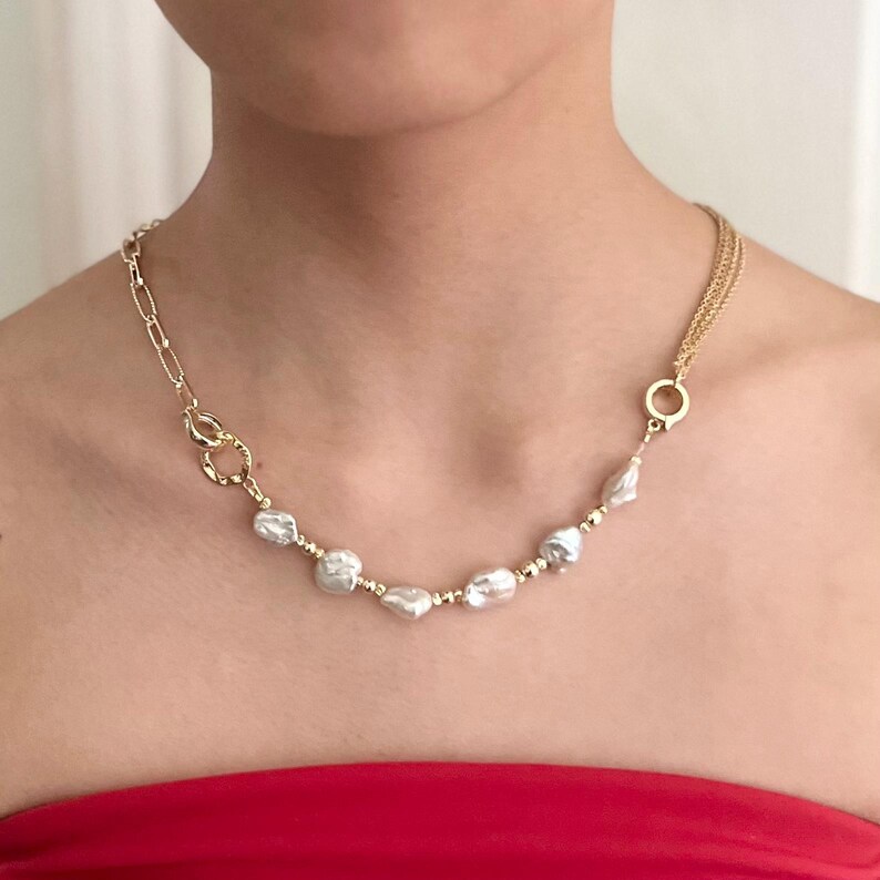 Peut inclure: Un collier dor&eacute; orn&eacute; de perles blanches irr&eacute;guli&egrave;res et de petites perles dor&eacute;es. Le collier a une cha&icirc;ne et une fermeture &agrave; fermoir d'un c&ocirc;t&eacute; et une cha&icirc;ne &agrave; plusieurs brins de l'autre. Le collier est port&eacute; sur un haut rouge.