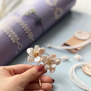 Puede incluir: Un delicado broche de flor blanca hecho de material nacarado con un centro dorado. El broche est&aacute; sujeto a una mano.