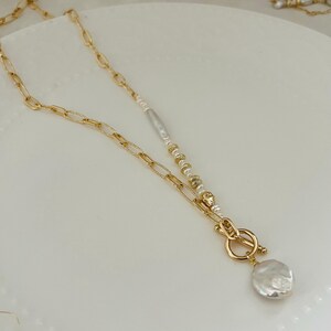 May include: A gold-coloured necklace with a toggle clasp and a large, irregular-shaped white pearl pendant. The necklace has a chain link design and a strand of small pearls and gold beads. The necklace is on a white surface.