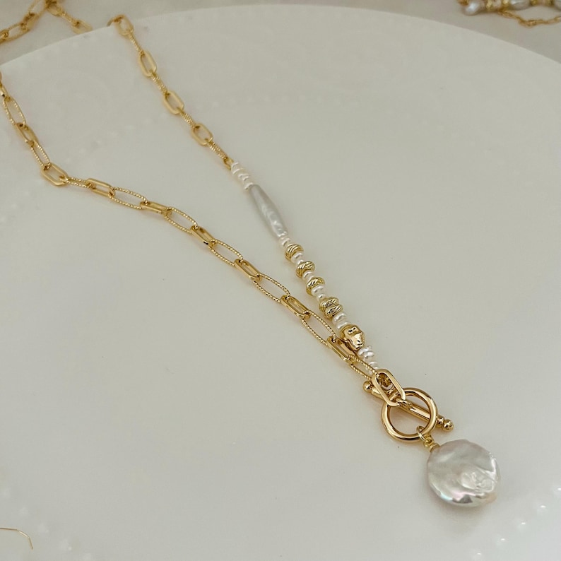 May include: A gold-coloured necklace with a toggle clasp and a large, irregular-shaped white pearl pendant. The necklace has a chain link design and a strand of small pearls and gold beads. The necklace is on a white surface.
