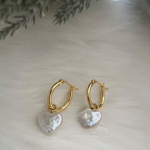 Op de afbeelding: Gouden creolen met een parelhartbedel. De oorbellen zijn van een eenvoudig en elegant ontwerp.