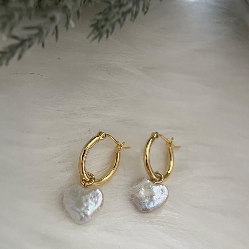 Op de afbeelding: Gouden creolen met een parelhartbedel. De oorbellen zijn van een eenvoudig en elegant ontwerp.