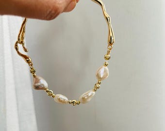 May include: A gold-coloured bracelet with an organic, wavy design. The bracelet features several irregularly shaped, off-white pearls interspersed with small, gold-coloured beads. The clasp is a hook and eye style.