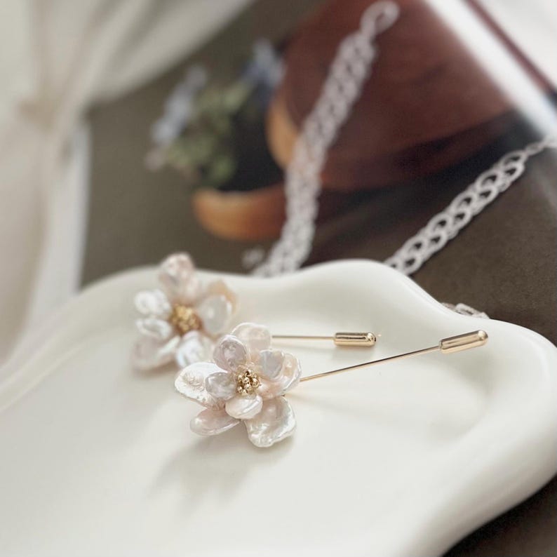 Puede incluir: Dos delicados broches de flores de perla con detalles dorados. Los broches est&aacute;n sobre una superficie blanca y est&aacute;n listos para ser usados.