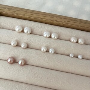 Peut inclure: Plusieurs paires de boucles d'oreilles en perles de diff&eacute;rentes tailles et couleurs, dont blanc et rose. Les boucles d'oreilles sont dispos&eacute;es sur une surface beige clair textur&eacute;e, dans un pr&eacute;sentoir en bois.