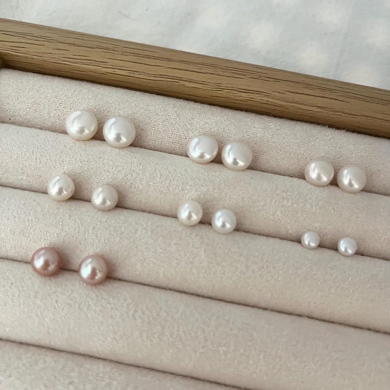 Peut inclure: Plusieurs paires de boucles d'oreilles en perles de diff&eacute;rentes tailles et couleurs, dont blanc et rose. Les boucles d'oreilles sont dispos&eacute;es sur une surface beige clair textur&eacute;e, dans un pr&eacute;sentoir en bois.