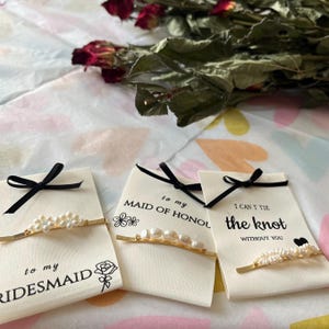 Puede incluir: Tres tarjetas con lazos negros y horquillas doradas adornadas con perlas. Las tarjetas dicen "to my bridesmaid", "to my maid of honour" y "I can't tie the knot without you". Rosas secas en el fondo.