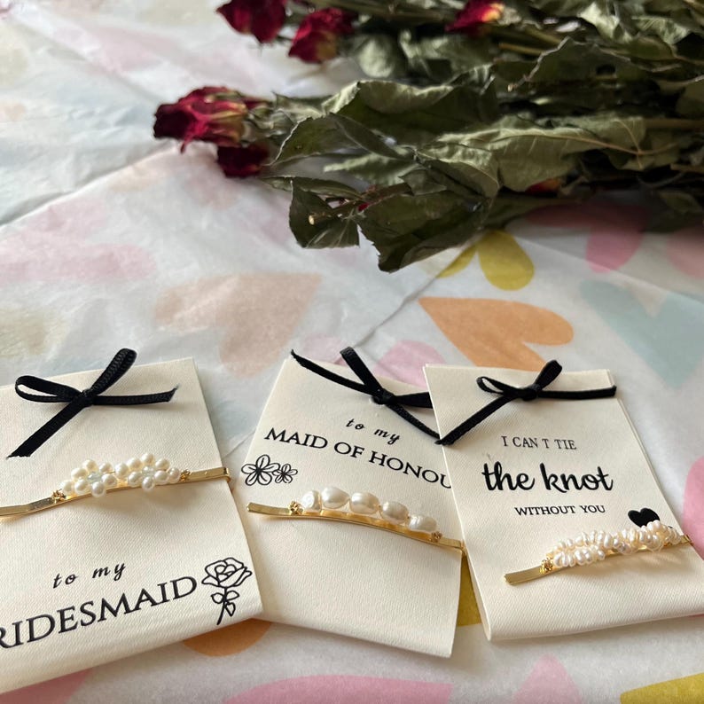 Puede incluir: Tres tarjetas con lazos negros y horquillas doradas adornadas con perlas. Las tarjetas dicen "to my bridesmaid", "to my maid of honour" y "I can't tie the knot without you". Rosas secas en el fondo.