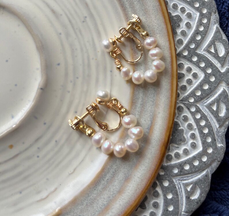 May include: A pair of gold-tone clip-on earrings with a row of white pearls around the edge of each earring.
