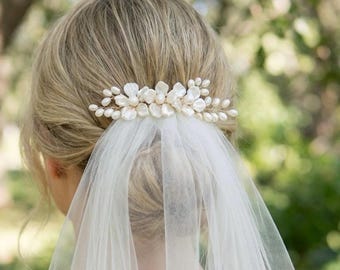 May include: A bridal hair comb featuring a cluster of white flower and pearl accents. The comb is set against a backdrop of blonde hair and a sheer white veil, creating an elegant and romantic look.