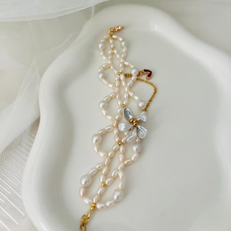 May include: A delicate pearl necklace with a floral accent. The necklace features a mix of small and large pearls, connected by gold-colored metal. A flower-shaped charm with white petals and a gold center adds a touch of elegance. The necklace is displayed on a white surface.