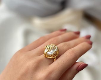 May include: A gold ring with a large white pearl set in a delicate, intricate design. The pearl is surrounded by a gold cage with small, clear stones.