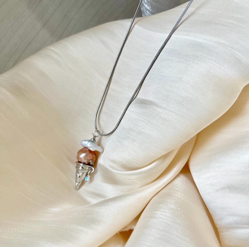 Peut inclure: Un collier en cha&icirc;ne argent&eacute;e avec un pendentif en forme de cornet de glace. Le cornet est rempli de trois boules de glace nacr&eacute;es dans des tons de rose et de blanc.