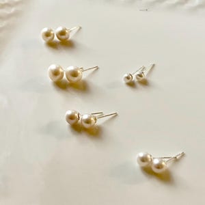 Peut inclure: Plusieurs paires de boucles d'oreilles en perles de diff&eacute;rentes tailles sont dispos&eacute;es sur une surface blanche. Les boucles d'oreilles ont des perles rondes classiques et des tiges en argent. Les perles sont de couleur cr&egrave;me.