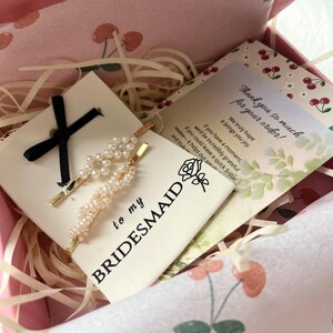 Puede incluir: Una caja de regalo que contiene dos horquillas de perlas, una cinta negra y una tarjeta que dice "to my bridesmaid". La caja tambi&eacute;n incluye una tarjeta de agradecimiento con un patr&oacute;n de flor de cerezo.