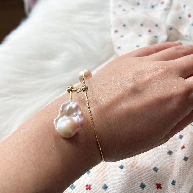 Peut inclure: Un bracelet en cha&icirc;ne dor&eacute;e avec un grand pendentif de perle irr&eacute;guli&egrave;re. La perle est de couleur blanc cass&eacute;.