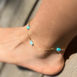 May include: A gold anklet with two chains and turquoise beads. The anklet is on a person's ankle.
