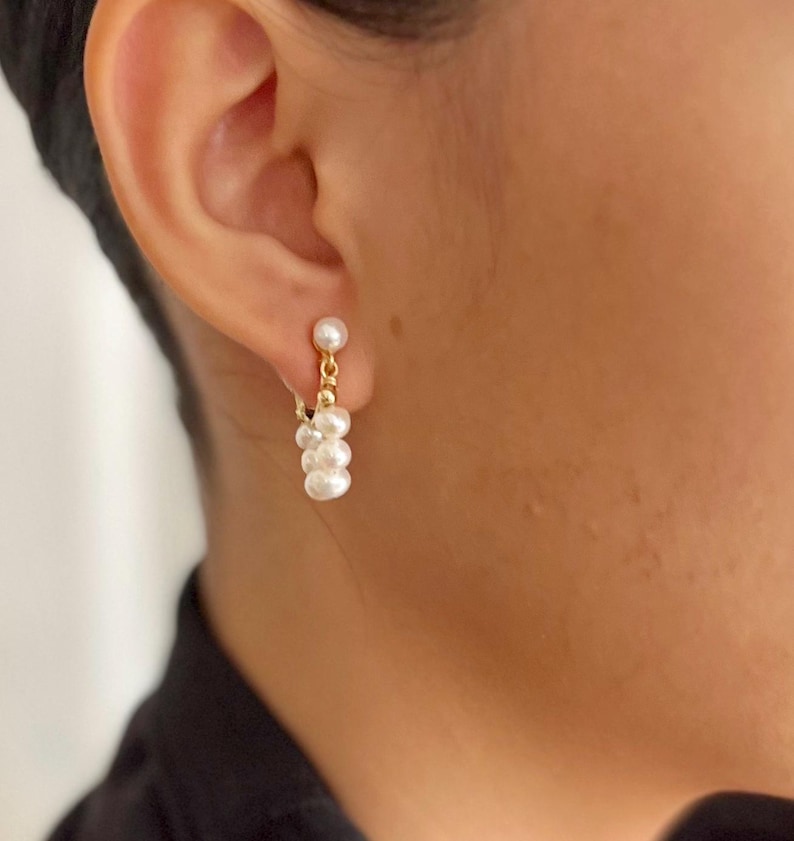 May include: A pair of gold hoop earrings with a cluster of white pearls dangling from each hoop.