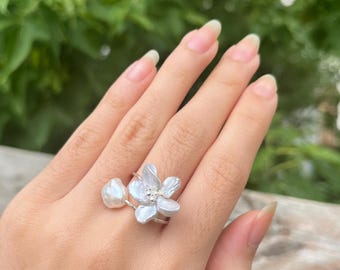 May include: A silver-toned ring featuring a white, iridescent, organic-shaped pearl flower design with five petals and a single, smaller, rounded white pearl accent attached to the side of the thin, polished metal band.