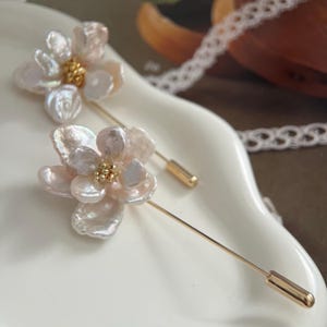 Puede incluir: Dos delicados broches de flores de perla con detalles dorados. Cada broche presenta un grupo de perlas blancas dispuestas en forma de flor con un centro dorado.